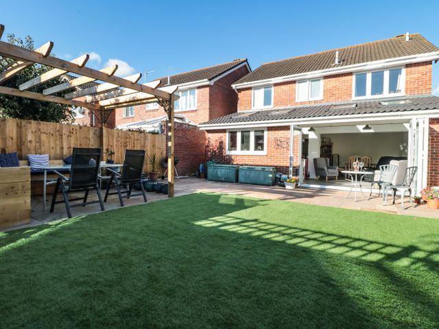 A backyard with artificial grass a wooden pergola with chairs and a table and a patio area leading into a house at Sandy Toes in Mudeford near Christchurch