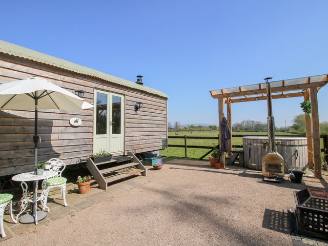 A wooden shepherd's hut with steps leading to a door beside a table with green cushioned chairs and umbrella with a hot tub under a wooden pergola in a fenced yard at Balwen Shepherd's Hut in Berriew