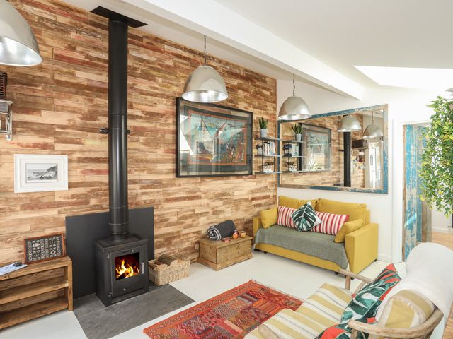 A living room with a wood stove yellow sofa wooden chair and a colorful rug at Lle Te in Abersoch