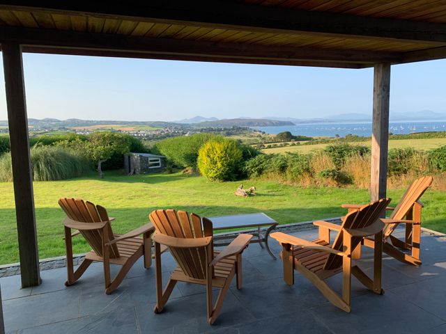 A covered patio with four wooden chairs and a glass table overlooking a grassy lawn and coastal landscape at Morawelon in Abersoch