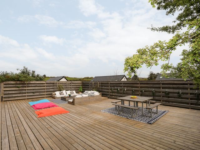 An outdoor wooden deck with a corner sofa area and colorful bean bags alongside a picnic table on a patterned rug at Nyth Y Fran in Abersoch