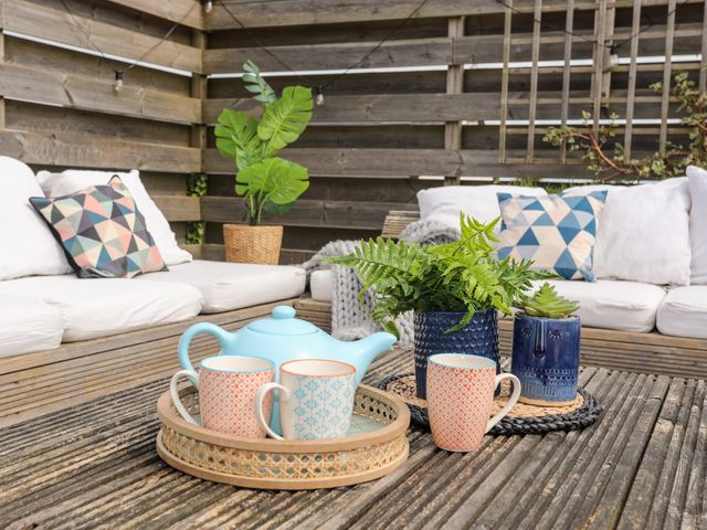 An outdoor seating area with white cushions colorful patterned pillows a teapot three mugs and two potted plants on a wooden table at Nyth Y Fran in Abersoch