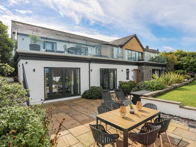 A patio area with a wooden dining table and chairs and reclining chairs outside a two-story house with glass balcony railings at Trem Y Borth Apartment in Bwlchtocyn near Abersoch