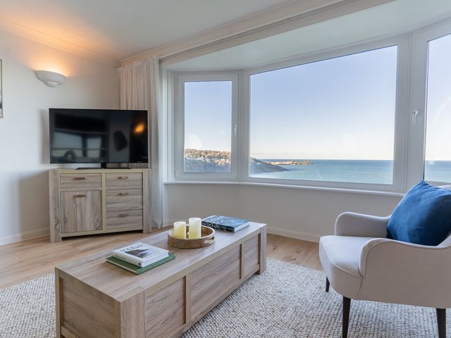 A living room with a TV on a wooden cabinet a wooden coffee table with books and candles a chair with a blue cushion and large windows with an ocean view at Flat 17 The Stones in Carbis Bay