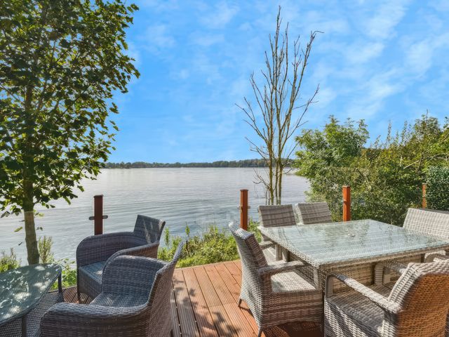 An outdoor deck with wicker chairs and glass tables overlooking a lake with trees at Gerrida Lodge South Lakeland Leisure Village near Tewitfield