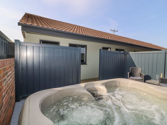 An outdoor patio area with a hot tub a chair a table with glasses and a bottle behind a grey fence at Chalet 1 in Sherburn
