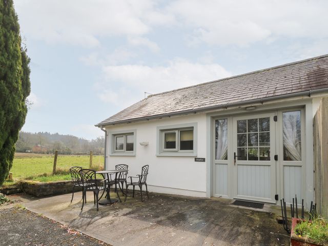 An outdoor patio area with a table and four metal chairs beside a single-story building with two small windows and a glass door at Dinefwr Cottage in Golden Grove near Llandeilo