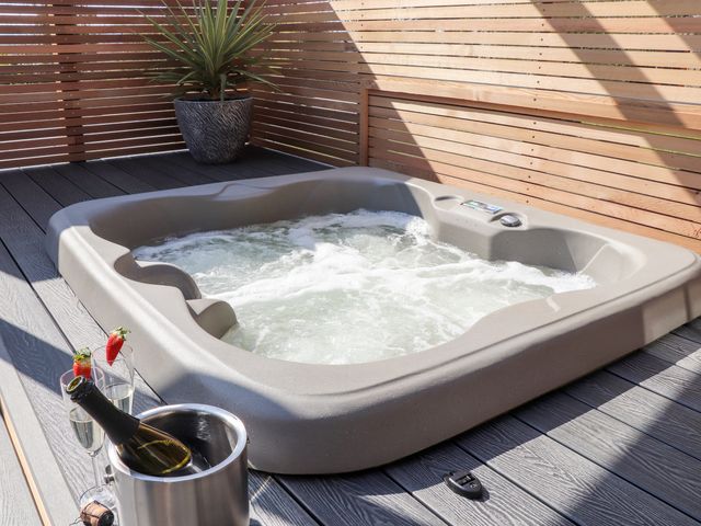 An outdoor hot tub on a wooden deck with a potted plant and two glasses of champagne with strawberries at Rosewarrick Cottage in Lanivet