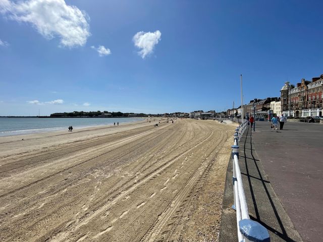 A sandy beach with tire tracks and footprints beside a paved promenade with pedestrians and buildings at Sunrise Apartment in Weymouth
