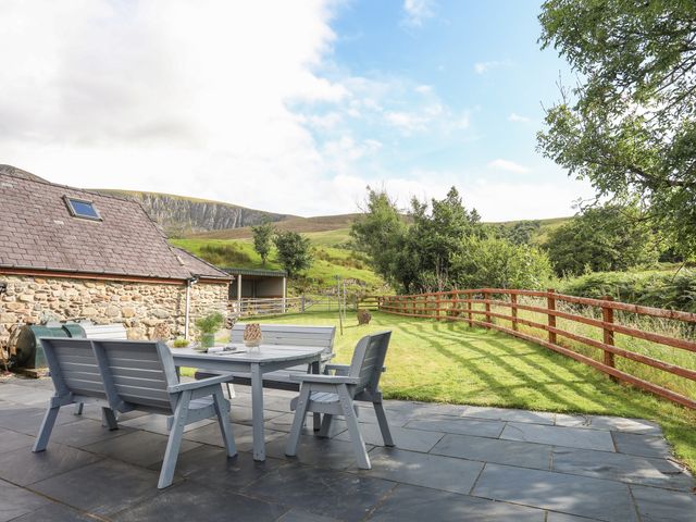 An outdoor patio with a table and chairs next to a stone building and a fenced grassy area with hills in the background at Ty Coch in Betws Garmon near Caernarfon