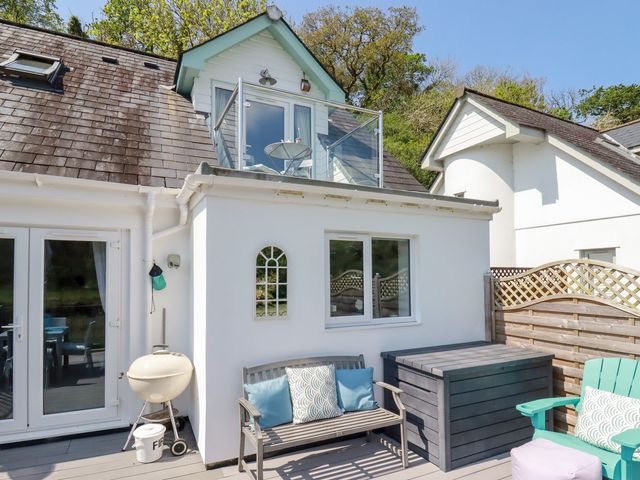 An outdoor patio area with a bench, cushions, a barbecue grill, a wooden storage box, a turquoise chair, and a small balcony above at Penpoll in Little Petherick Creek near Padstow
