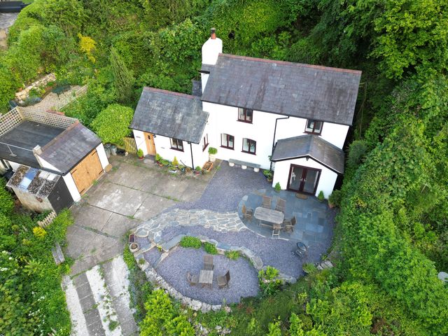 A white house with slate roof surrounded by dense greenery with outdoor seating areas and a driveway at Alwyn Cottage in Froncysyllte