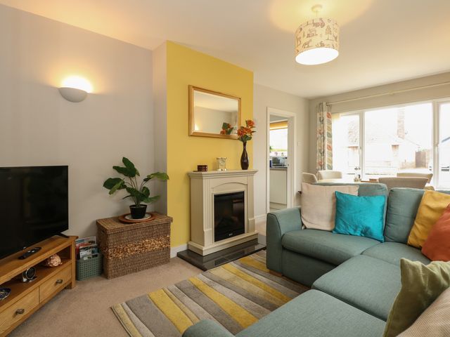 A living room with a green sectional sofa and colorful cushions a striped rug a fireplace with a mirror above and a television on a wooden stand at The Lobster Pot in Overstrand