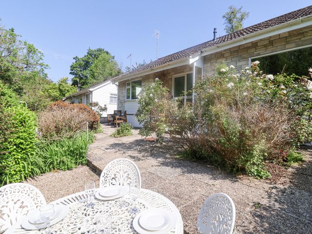 An outdoor patio area with a white metal table and chairs set with plates and glasses next to a stone house with flowering bushes at High Trees in Lyme Regis