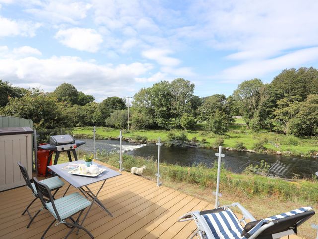 An outdoor deck with a table and chairs a barbecue and a striped lounge chair overlooking a river and trees at Llety'r Bugail 1 in Llanrug