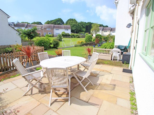 An outdoor patio with a white table and six chairs overlooking a garden with plants and houses in the background at Green Cottage in Borth-Y-Gest