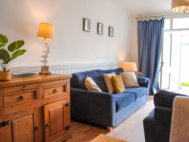 A living room with a blue sofa and cushions wooden cabinet with lamp and plant blue curtains and an open door at Paddler's Rest in Hope Cove