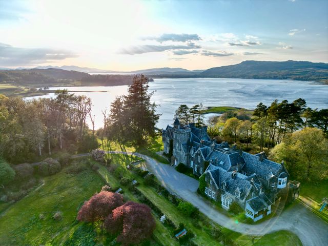An aerial view of a large stone house with multiple roofs near a body of water surrounded by trees and hills at Achnacloich in Connel