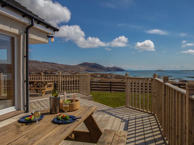 A wooden deck with a picnic table set with plates and glasses overlooking a rocky shoreline and sea at Rockpool in Glendale Isle of South Uist