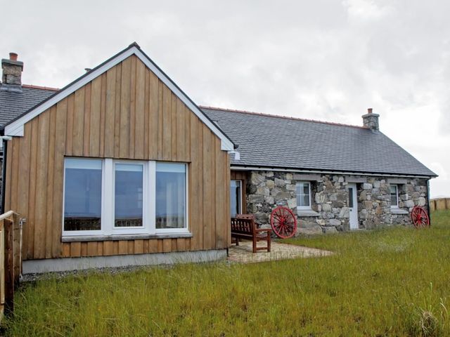 A house with wooden and stone walls a bench and red wagon wheels on the grass at Beach View Cottage in Daliburgh