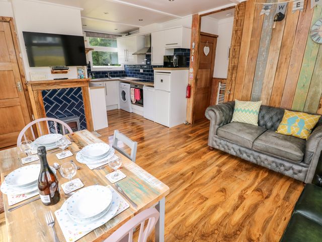 An open kitchen and dining area with a wooden table set with plates and glasses a sofa with cushions a wall-mounted tv and wooden paneling on the wall at Ty Haf in Roch