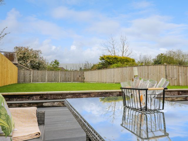 A backyard with green grass and wooden fencing with a glass table holding a basket of glasses and a bottle at 2 Penporth in Rhayader