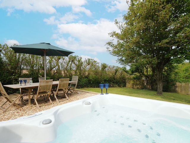 An outdoor patio with a wooden dining table and chairs under an umbrella next to a hot tub in a garden at Carvannel Cottages North Cliffs near Portreath