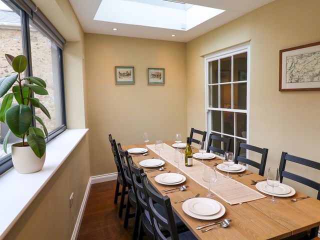 A dining room with a wooden table set for eight people a potted plant by the window and framed pictures on the walls at The Fernery in Colyton