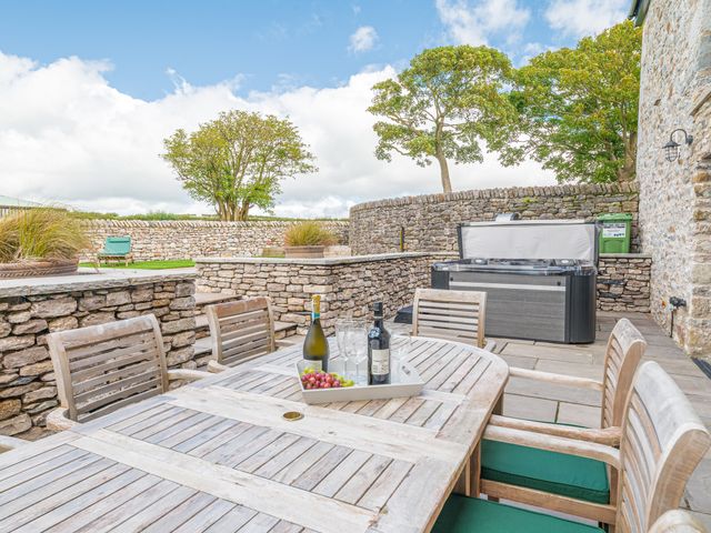 An outdoor patio with a wooden table and chairs a wine bottle and glasses on the table a hot tub and stone walls at Old Stable in Roose