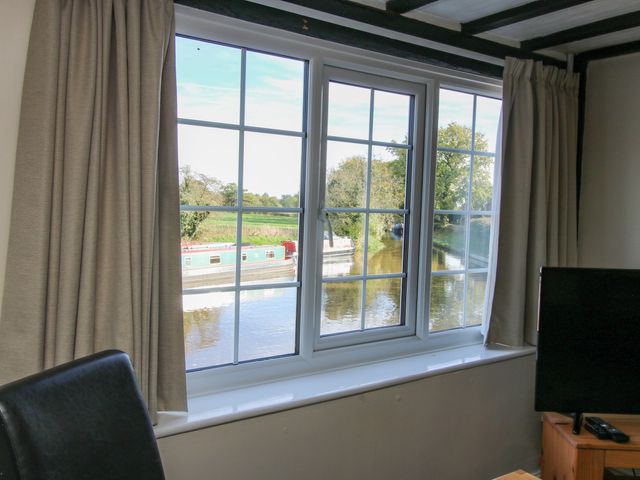 A window with beige curtains overlooking a canal with narrowboats visible from inside a room at Mill End in Wrenbury