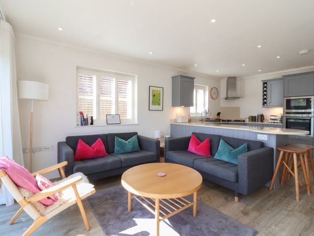 A living room with two gray sofas, a round wooden coffee table, a wooden armchair with pink cushions and an open kitchen with gray cabinets at Crabapple Cottage Southwold