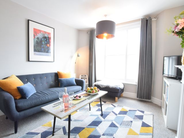 A living room with a blue sofa orange and blue cushions a coffee table with a bottle and glasses patterned rug and a window with grey curtains at Driftwood Southwold