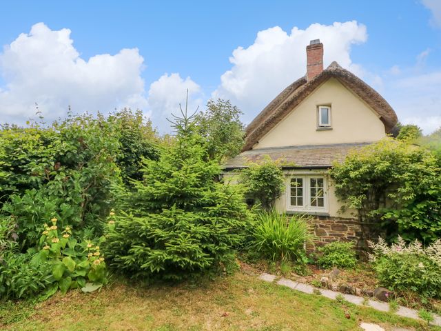 A thatched roof cottage with a stone base surrounded by greenery and a small pathway at Rose Cottage in Okehampton