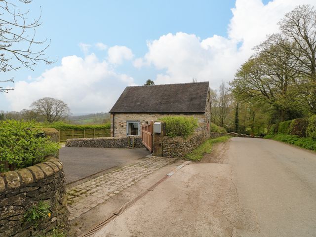 A stone cottage with a wooden gate by a paved road surrounded by trees and stone walls at Postbox Cottage in Atlow near Ashbourne