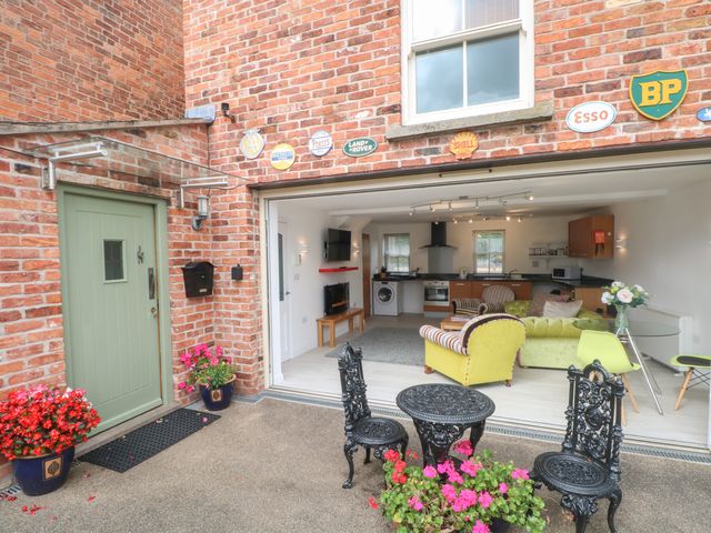 An outdoor patio with black metal table and chairs and pink flowers in pots next to a green door and an open living room with kitchen at The Lodge at Cross Cottage in Holt North Wales