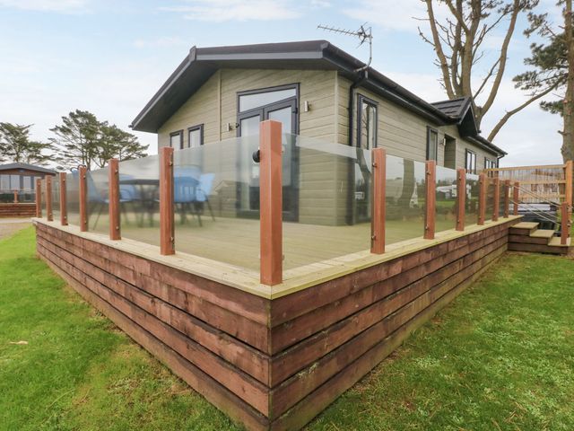 A raised wooden deck with glass railing around a small house on grass at Driftwood Lodge in Hasguard Cross near Broad Haven