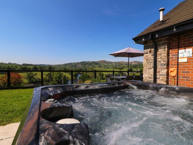 An outdoor hot tub next to a stone and brick building with a table, chairs, and umbrella overlooking green fields and hills at Pen Y Banc in Llanfaredd near Builth Wells