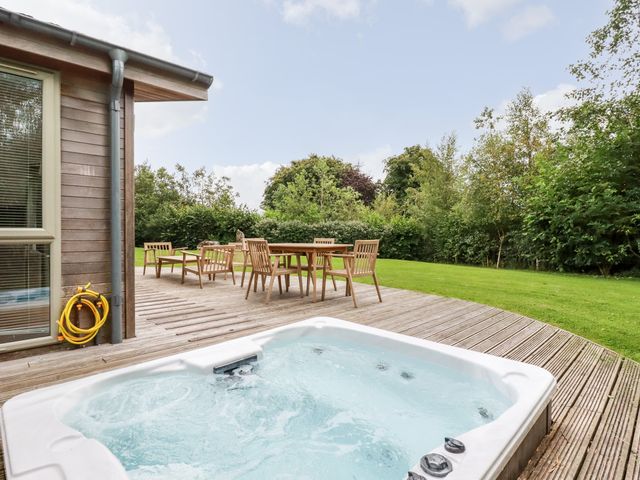 An outdoor area with a hot tub on a wooden deck and a wooden table with six chairs surrounded by grass and trees at 14 Horizon View in Dobwalls