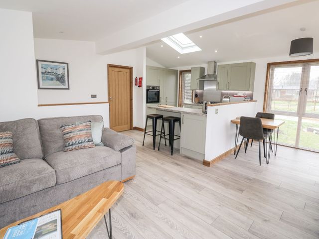 An open plan living room kitchen with a gray sofa wooden coffee table kitchen island with stools dining table with chairs and large windows at 2 Meadow Retreat in Dobwalls