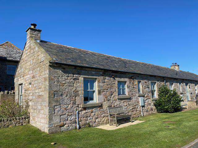 A stone cottage with windows a wooden bench and a small bush on a lawn at Beachward in Embleton