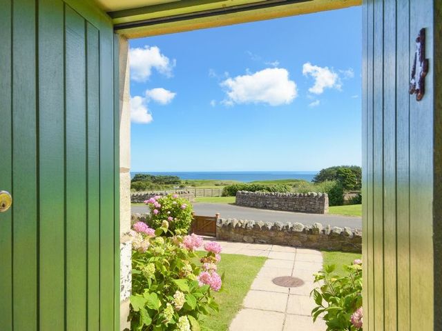 View through open green doors showing a stone path, garden with pink flowers, stone wall, grassy field, and ocean in the distance at Ottoline Cottage in Embleton