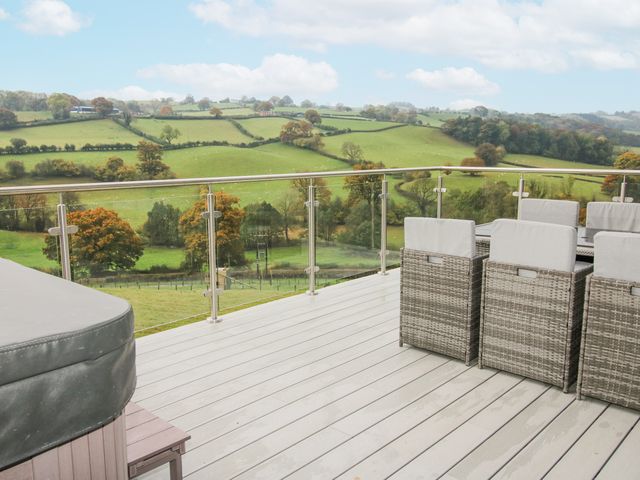 An outdoor deck with wicker chairs a table and glass railings overlooking green fields and trees at Bwncath in Garthmyl