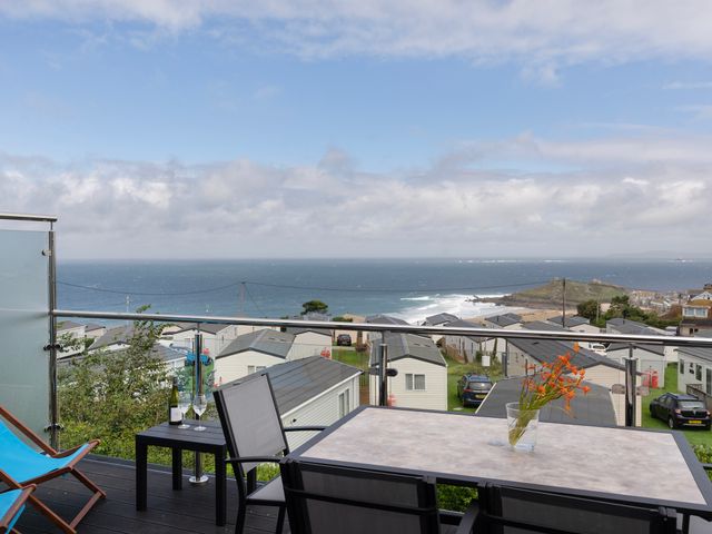 A balcony with a table, chairs, flowers, and a bottle of wine overlooking a caravan park and the sea at Treen in St Ives