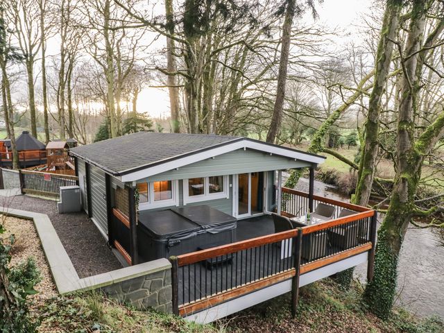 A cabin with a covered hot tub and outdoor seating on a deck overlooking a river surrounded by trees at Buttermilk Bridge in Dungiven