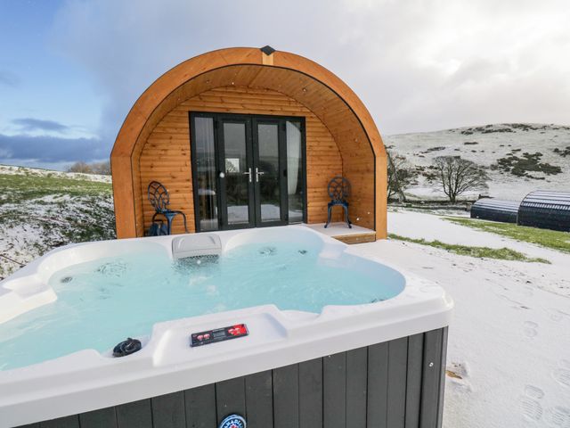 An outdoor hot tub in front of a wooden pod with black double doors and two chairs on a snowy hillside at Oak in Ulverston