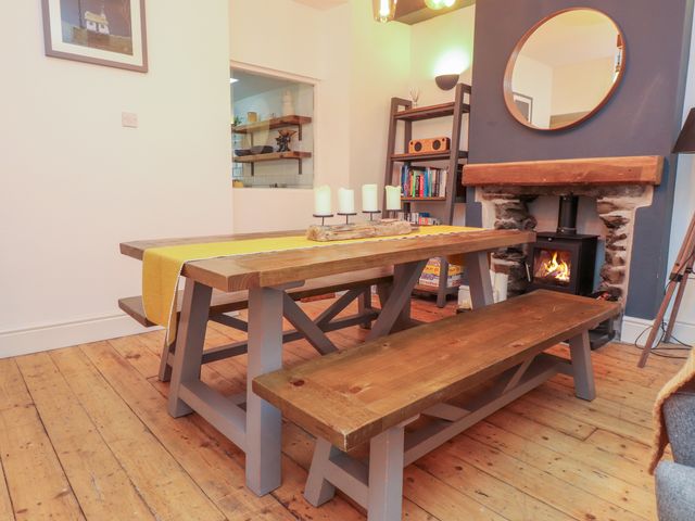 A dining area with a wooden table and benches a fireplace with a fire a round mirror and a bookshelf at Heol Madog in Porthmadog