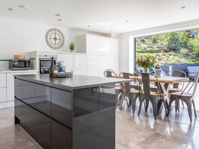 A kitchen with a large island, dining table with metal chairs, and a large open window showing a garden at 7 Beachdown in Challaborough