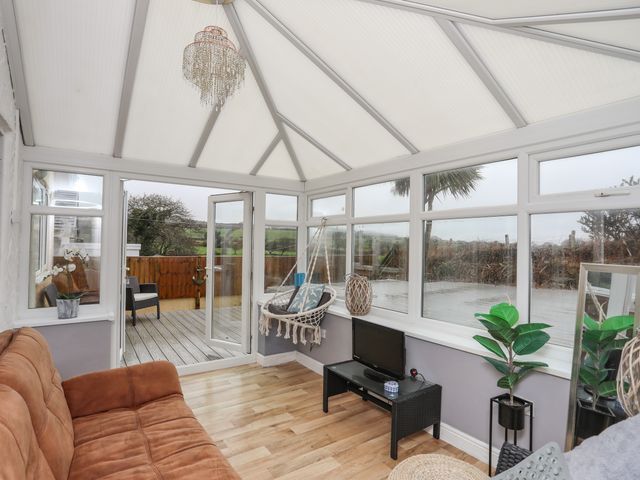 A conservatory with a brown sofa a hanging chair a small black TV stand with a TV a potted plant and a door leading to a wooden deck at Gwen Y Wawr in Llanbedrog