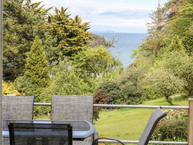 A garden with trees and a sea view behind outdoor chairs and a table at Flushing Meadow in Manaccan near Helford