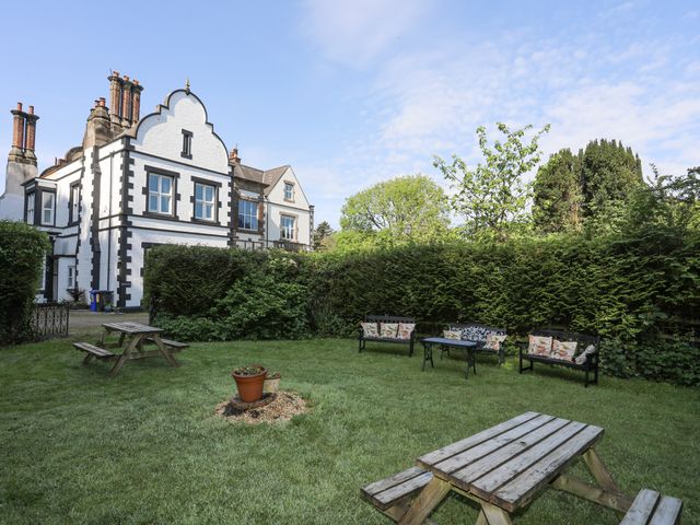 A garden area with two wooden picnic tables a metal seating set with cushions and a white house with chimneys in the background at Llys y Graig in Pentraeth